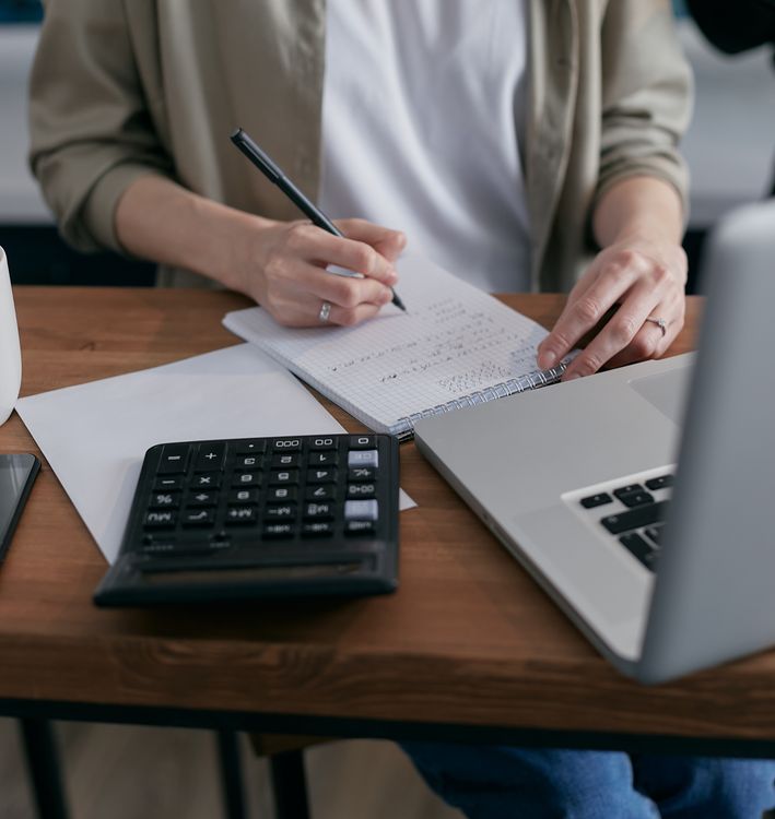 Frau berechnet Zahlen in einem Notizbuch mit Taschenrechner und Laptop – Stundensatz für Freelancer kalkulieren